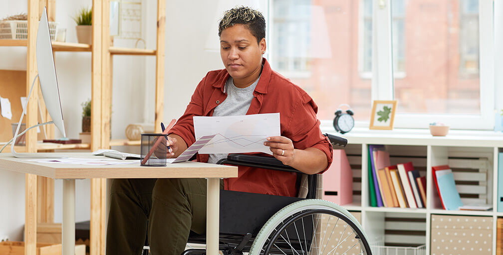 A woman in wheelchair sits at a desk writing and looking at papers. Apply for Supplemental Security Income in Oregon with help from our lawyers.