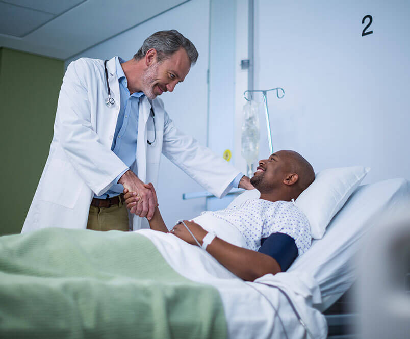 A man lies in a hospital bed, shaking hands with a doctor standing over him.